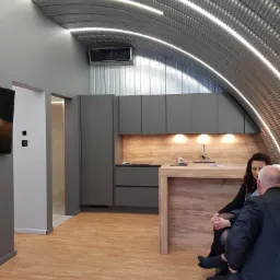 Wnętrze nowoczesnego domu z łukowym sufitem z blachy trapezowej, oświetlonym listwami LED, z zamontowaną klimatyzacją, szarą zabudową kuchenną i drewnianym blatem, gdzie kobieta i mężczyzna siedzą...