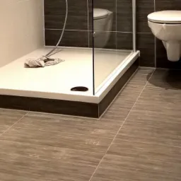 Wykończona łazienka z kabiną prysznicową, wiszącą toaletą i szarymi płytkami imitującymi drewno na podłodze i ścianach, widok na brodzik z odpływem i leżącą szmatką.