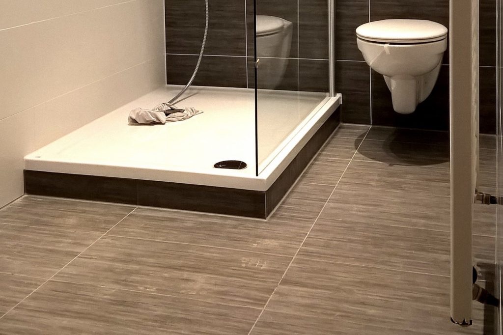 Wykończona łazienka z kabiną prysznicową, wiszącą toaletą i szarymi płytkami imitującymi drewno na podłodze i ścianach, widok na brodzik z odpływem i leżącą szmatką.