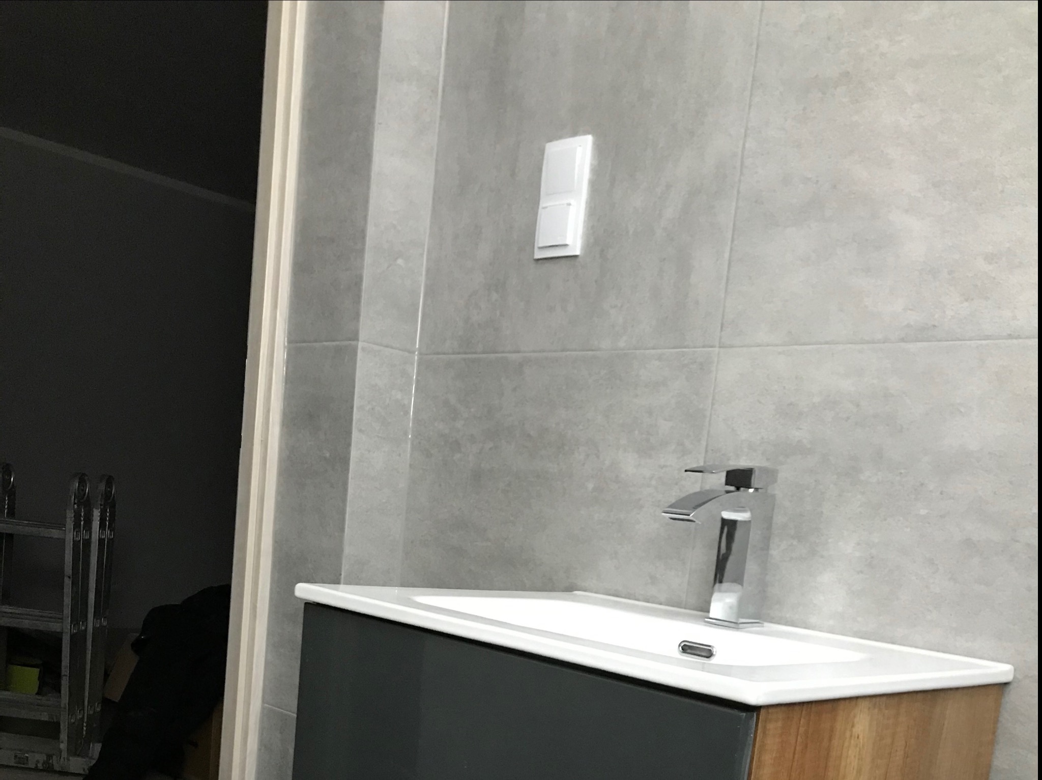 Szare płytki na ścianie łazienki z zamontowanym białym włącznikiem światła, umywalka z szafką w odcieniach szarości i drewna, widoczna chromowana bateria i drabina w tle.