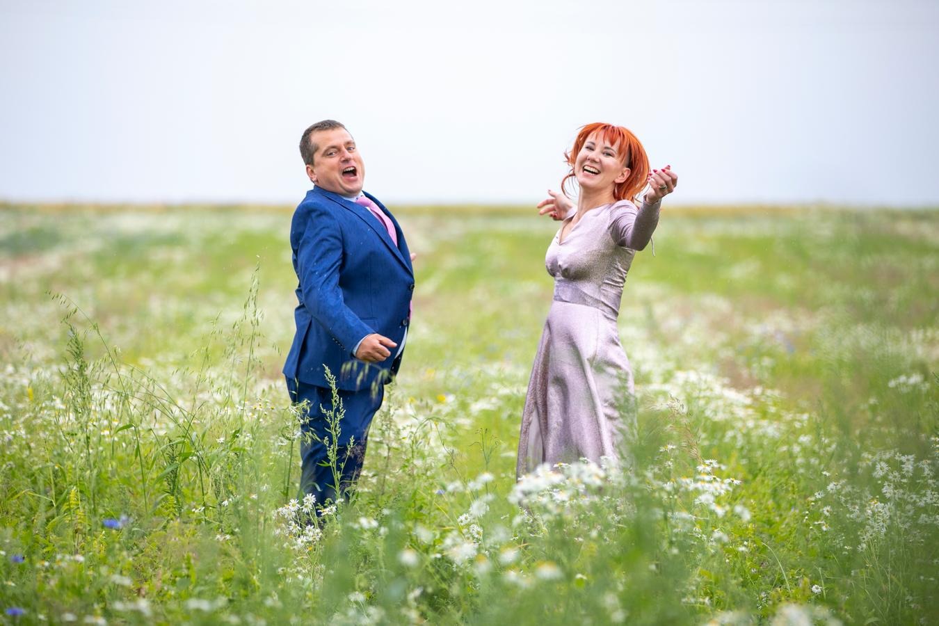 Para młoda w radosnych pozach na łące pełnej kwitnących rumianków, pan młody w niebieskim garniturze i różowym krawacie, panna młoda w srebrzystej sukni.