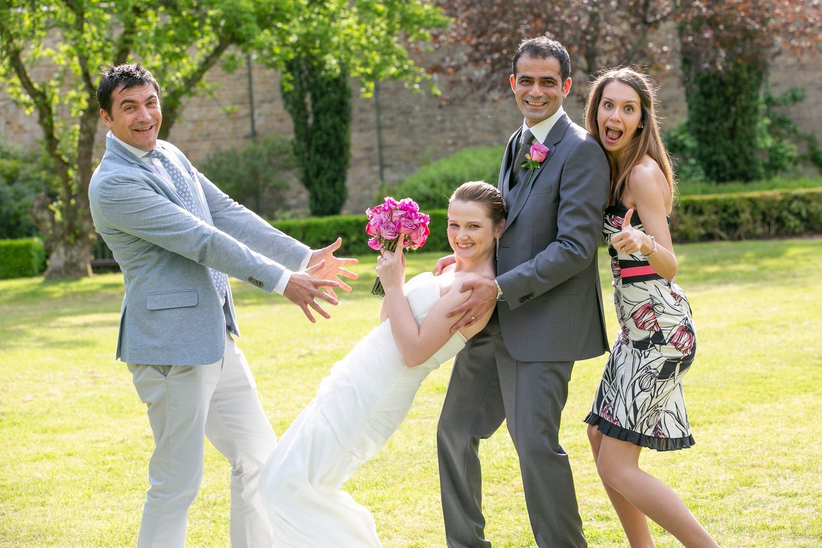Uśmiechnięta panna młoda w białej sukni, trzymająca bukiet różowych kwiatów, pozuje z uśmiechniętym panem młodym w szarym garniturze i dwoma gośćmi weselnymi na zielonym trawniku, jeden z gości...