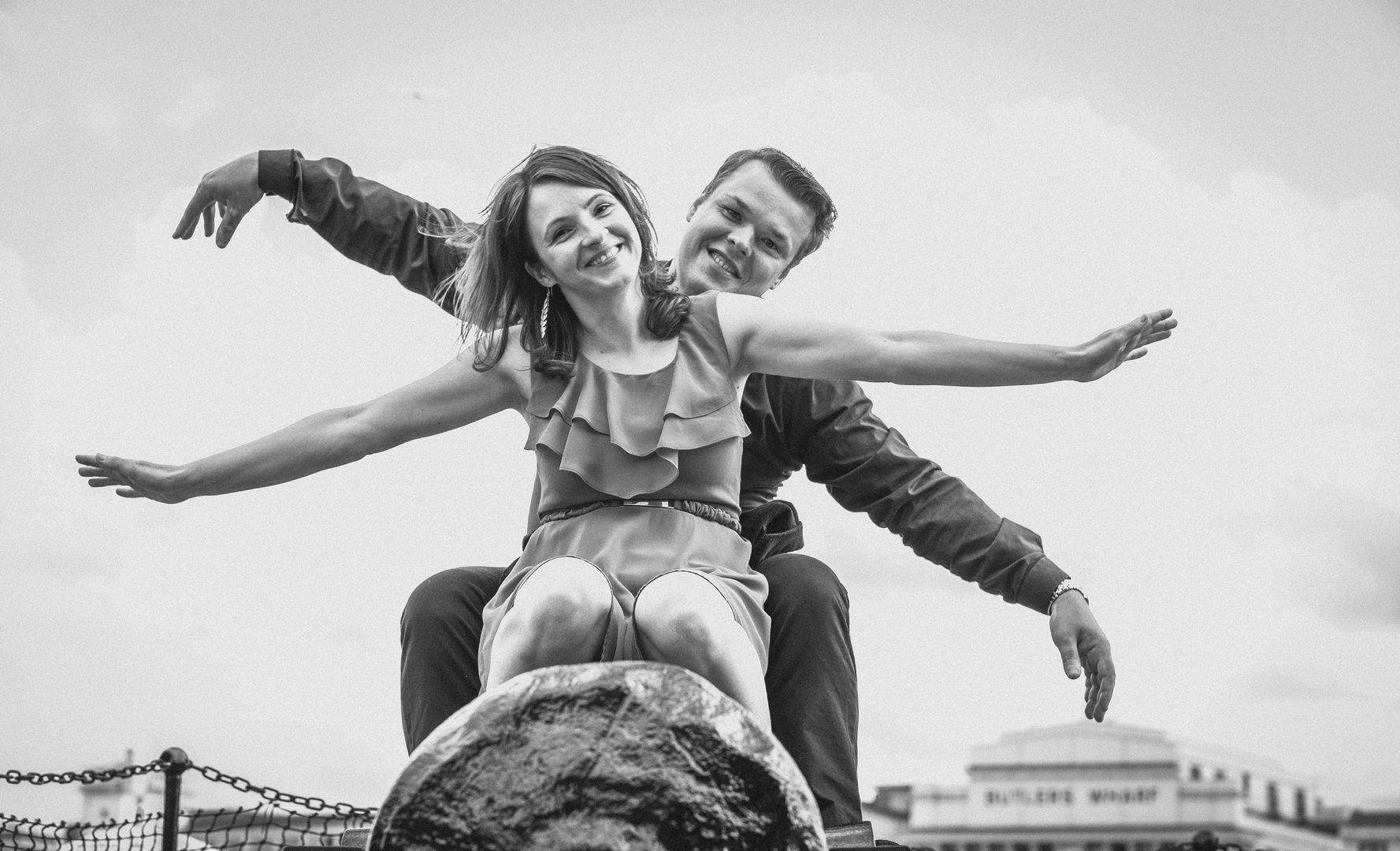 Czarno-białe zdjęcie uśmiechniętej pary na tle nieba i budynków. Kobieta siedzi na kamiennym cokole, a mężczyzna stoi za nią, oboje z rozłożonymi ramionami, imitując lot.