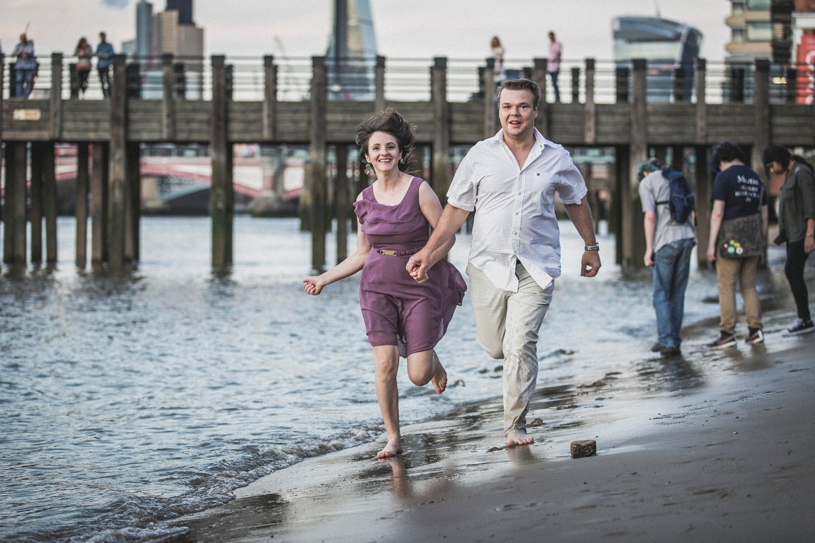 Para trzymająca się za ręce biegnie boso po mokrej plaży w kierunku widza, w tle drewniane molo i budynki miejskie.