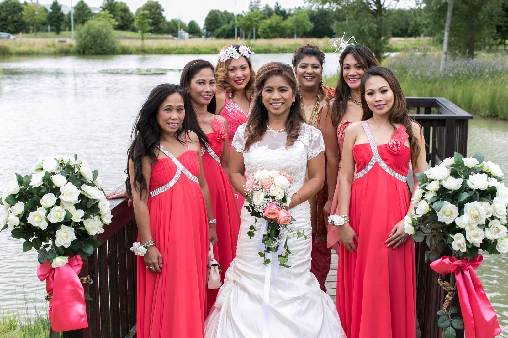 Panna młoda w białej sukni ślubnej z bukietem róż, otoczona druhnami w koralowych sukienkach, pozuje na drewnianym pomoście z widokiem na jezioro i zieleń w tle.