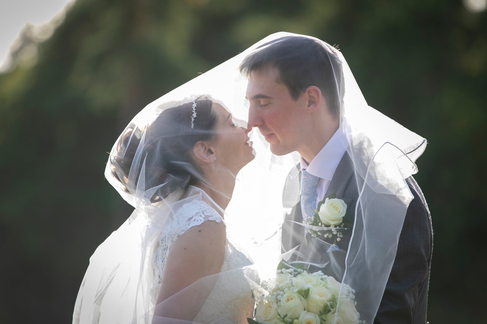 Para młoda, panna młoda w koronkowej sukni i diademie, pan młody w szarym garniturze z butonierką, stoją pod welonem, dotykając się nosami, w tle rozmyta zieleń.