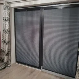 Dwa okna przesuwne z szarymi plisowanymi osłonami okiennymi, po bokach zwisają wzorzyste zasłony w odcieniach szarości i bieli.