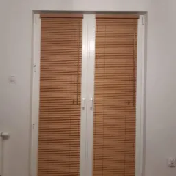 Drewniane żaluzje poziome zamontowane na dwuskrzydłowych drzwiach balkonowych, widok z wnętrza pomieszczenia.