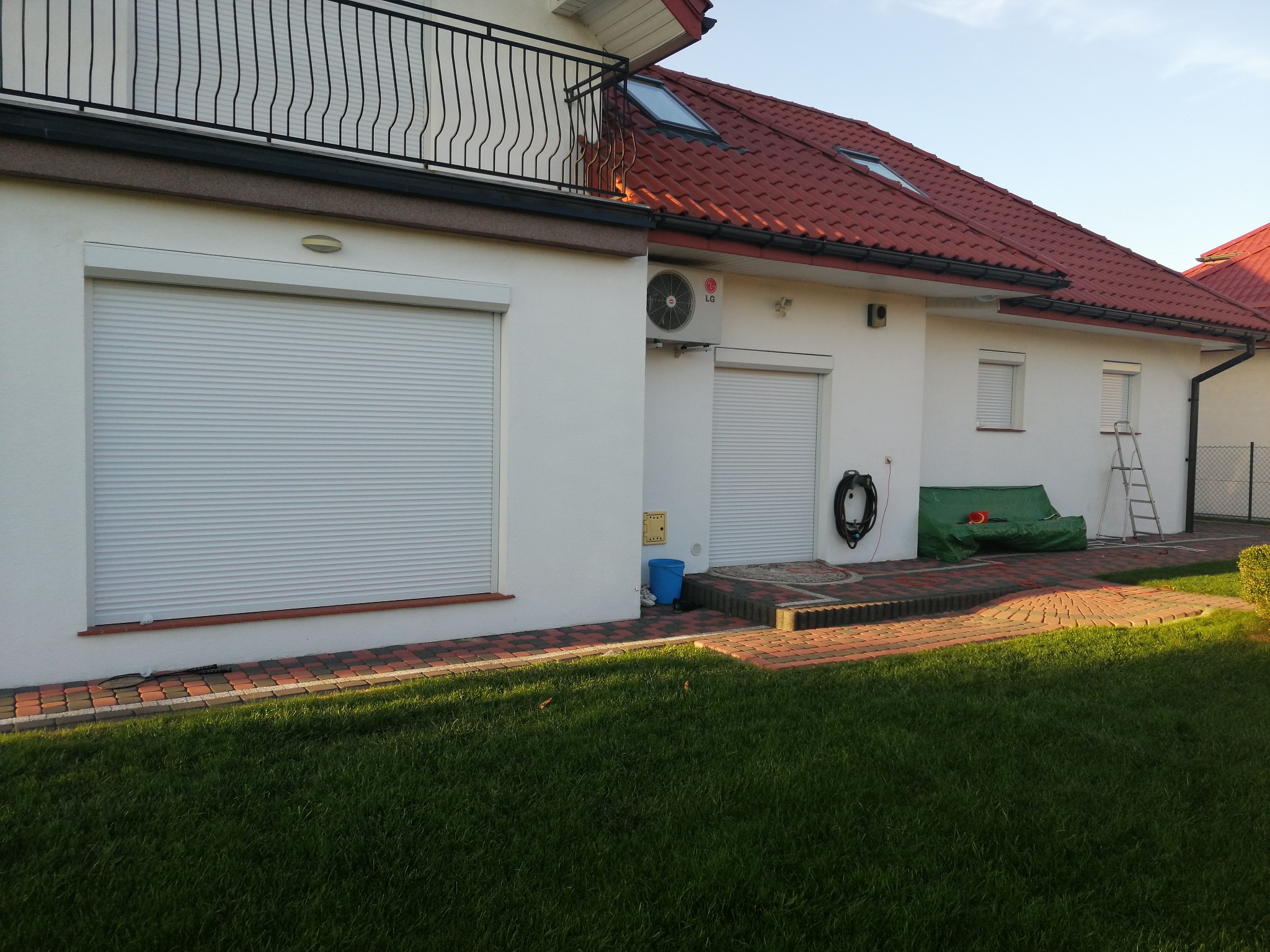 Biały dom jednorodzinny z czerwonym dachem i zamkniętymi, zewnętrznymi osłonami okiennymi, widok z trawnika w słoneczny dzień.