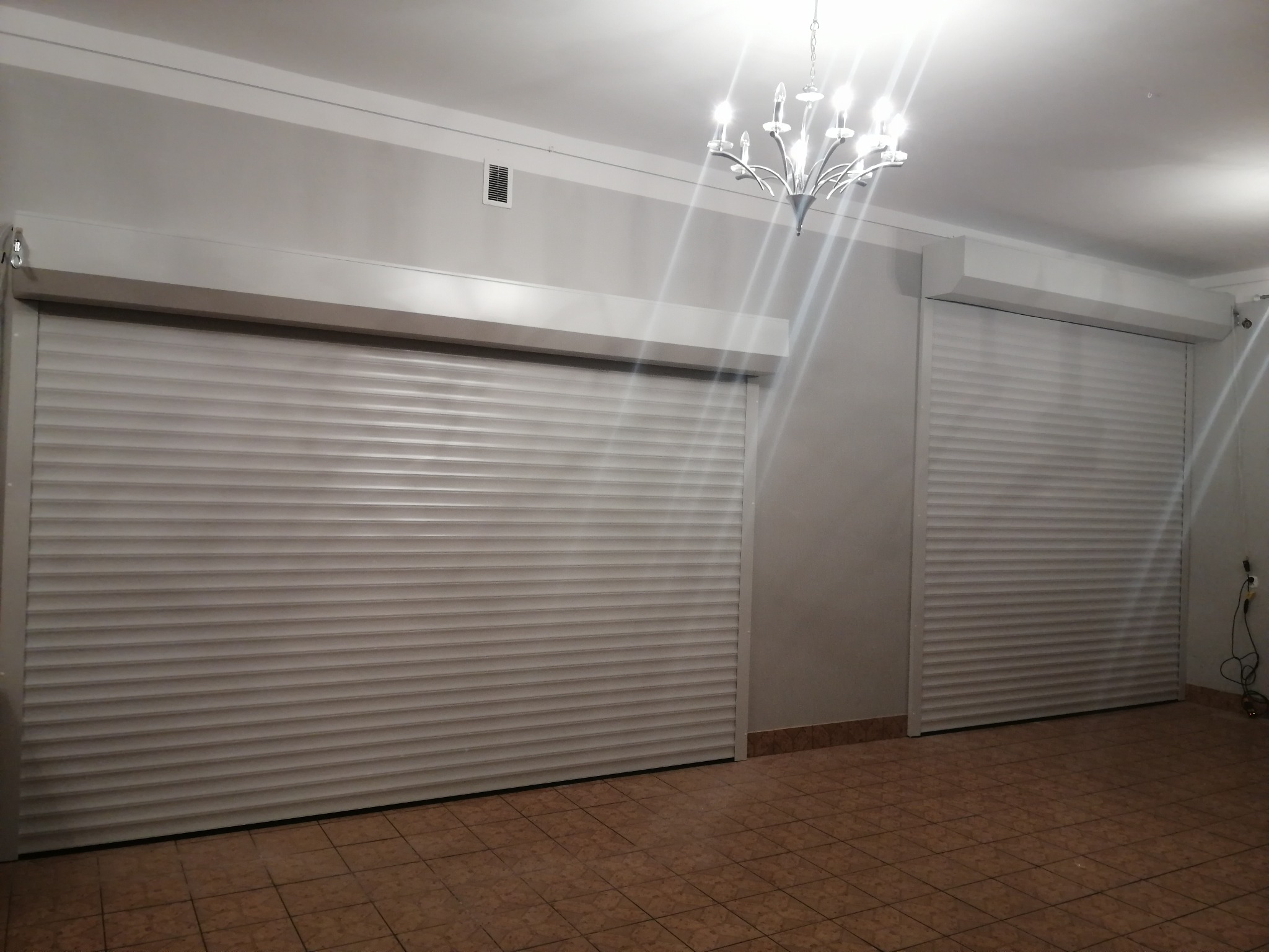 Dwa okna zabezpieczone zamkniętymi, jasnymi roletami w pomieszczeniu z brązową podłogą i kryształowym żyrandolem.