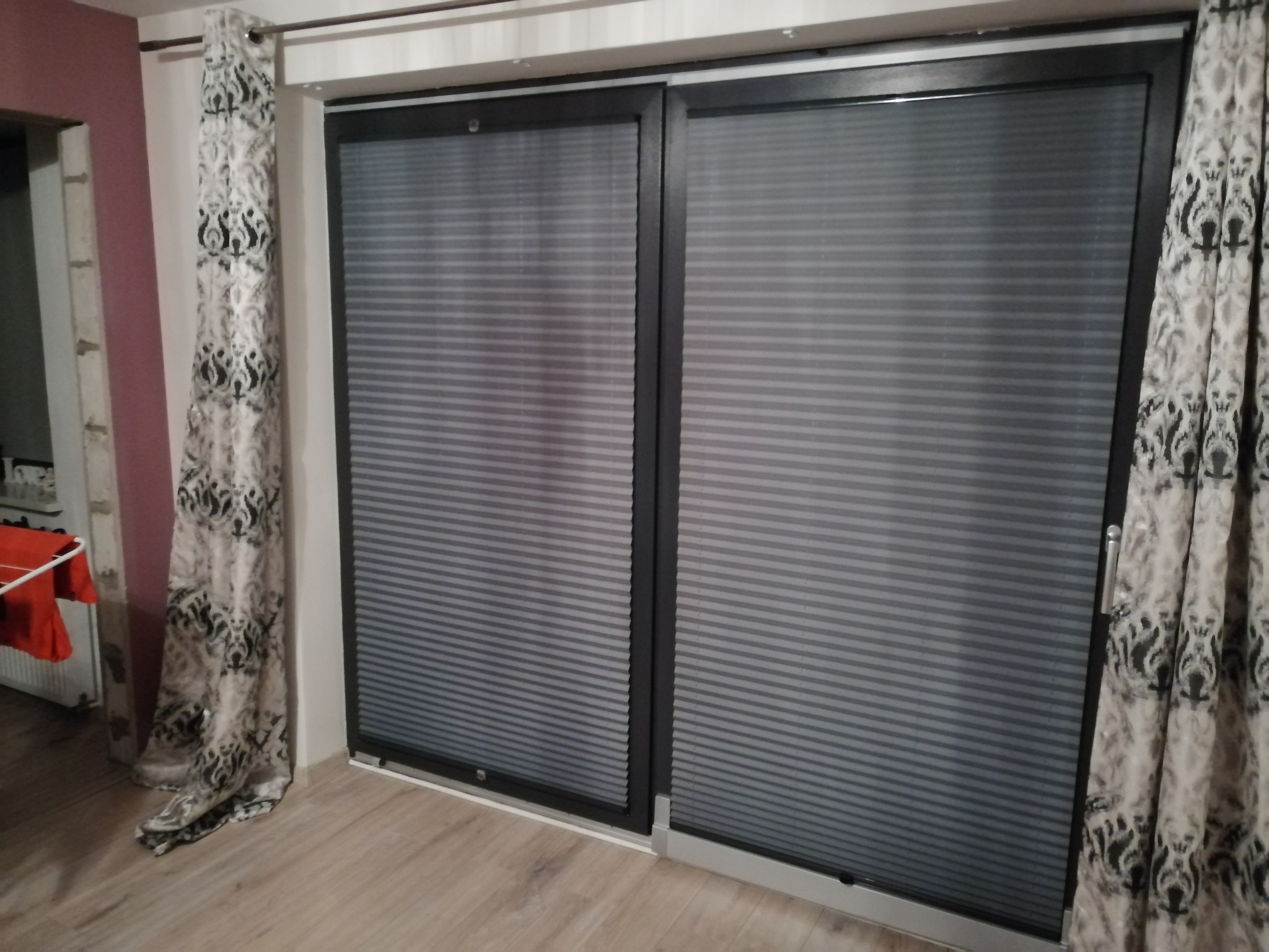 Dwa okna przesuwne z szarymi plisowanymi osłonami okiennymi, po bokach zwisają wzorzyste zasłony w odcieniach szarości i bieli.