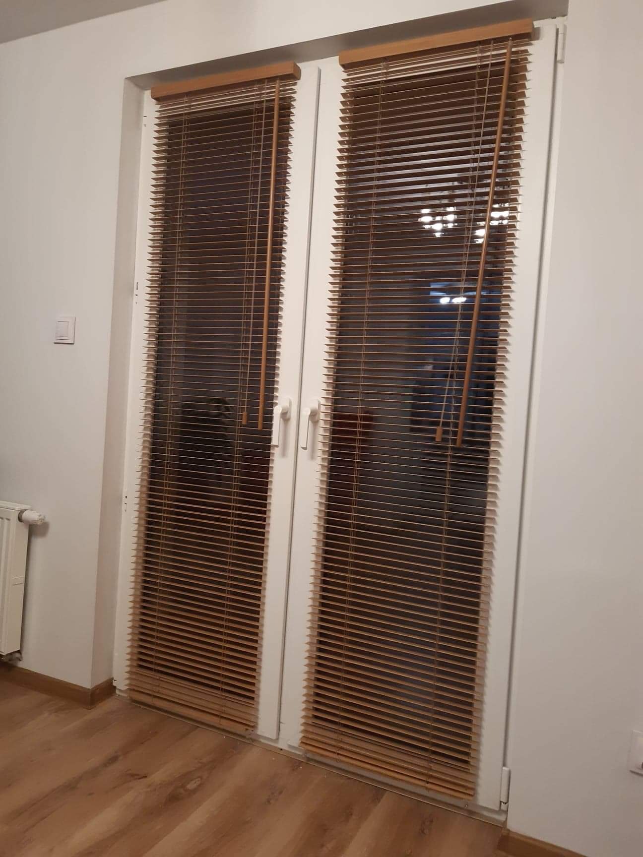 Drewniane żaluzje poziome zamontowane na białych drzwiach balkonowych, widoczne z perspektywy wnętrza pomieszczenia, z fragmentem grzejnika i podłogi imitującej drewno.
