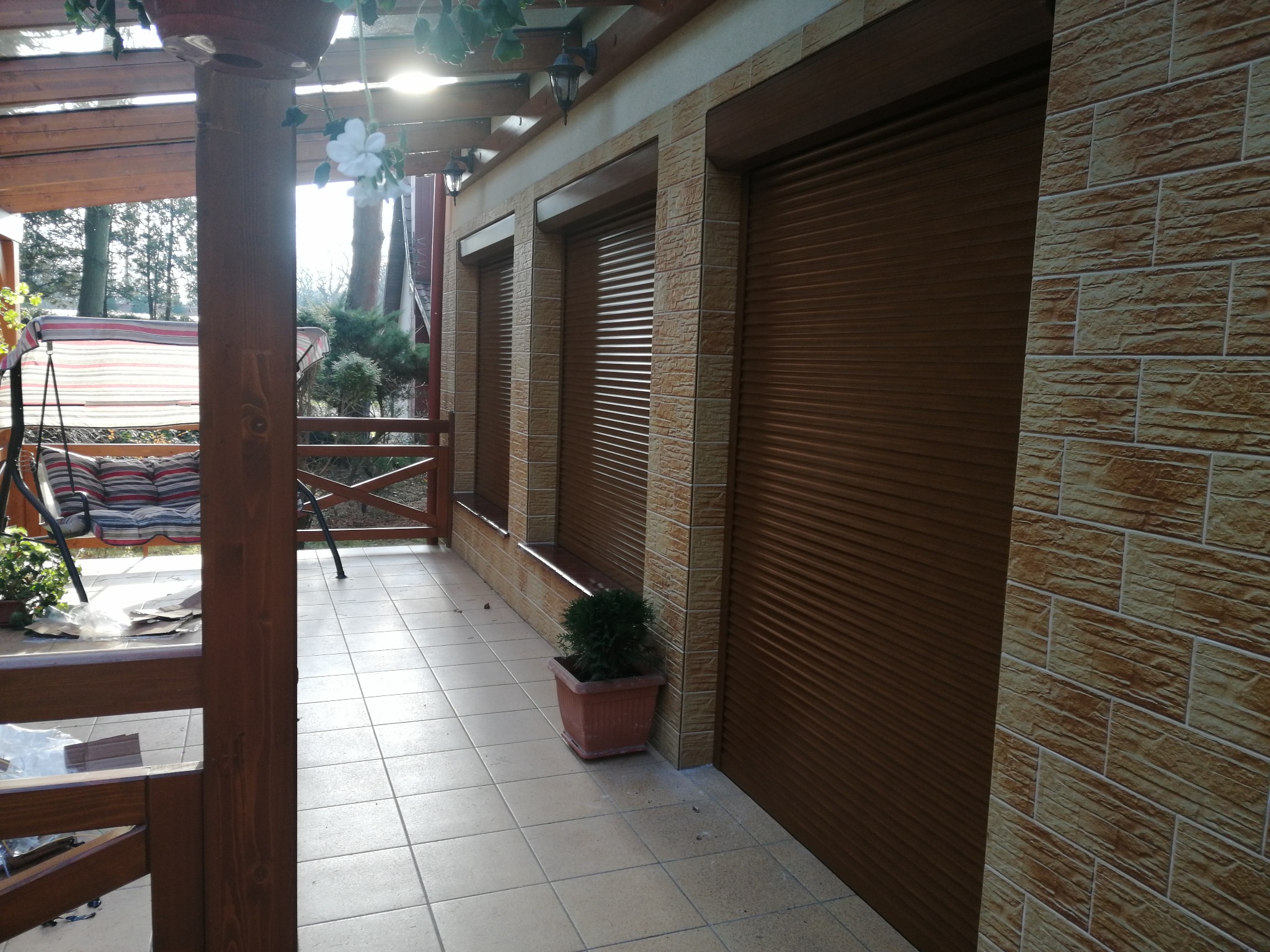 Zewnętrzne brązowe rolety zamontowane na elewacji budynku z cegieł, widok z zadaszonego tarasu z drewnianą konstrukcją.