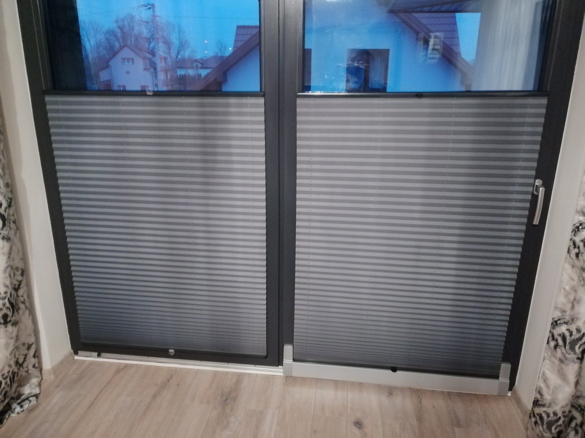 Szare plisy okienne zamontowane w oknie balkonowym, widok na zewnątrz z domem i drzewami.