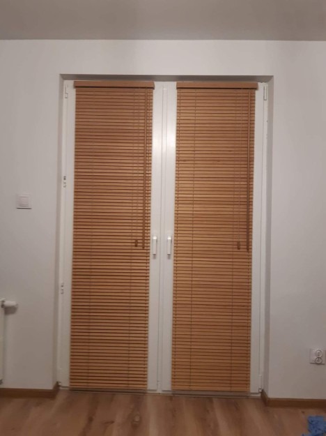 Drewniane żaluzje poziome zamontowane na dwuskrzydłowych drzwiach balkonowych, widok z wnętrza pomieszczenia.