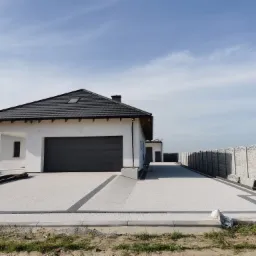 Nowoczesny dom jednorodzinny z jasną elewacją i czarnym dachem, podjazd wyłożony jasną kostką brukową z ciemnymi akcentami, widoczne studzienki odpływowe i fragment ogrodzenia.