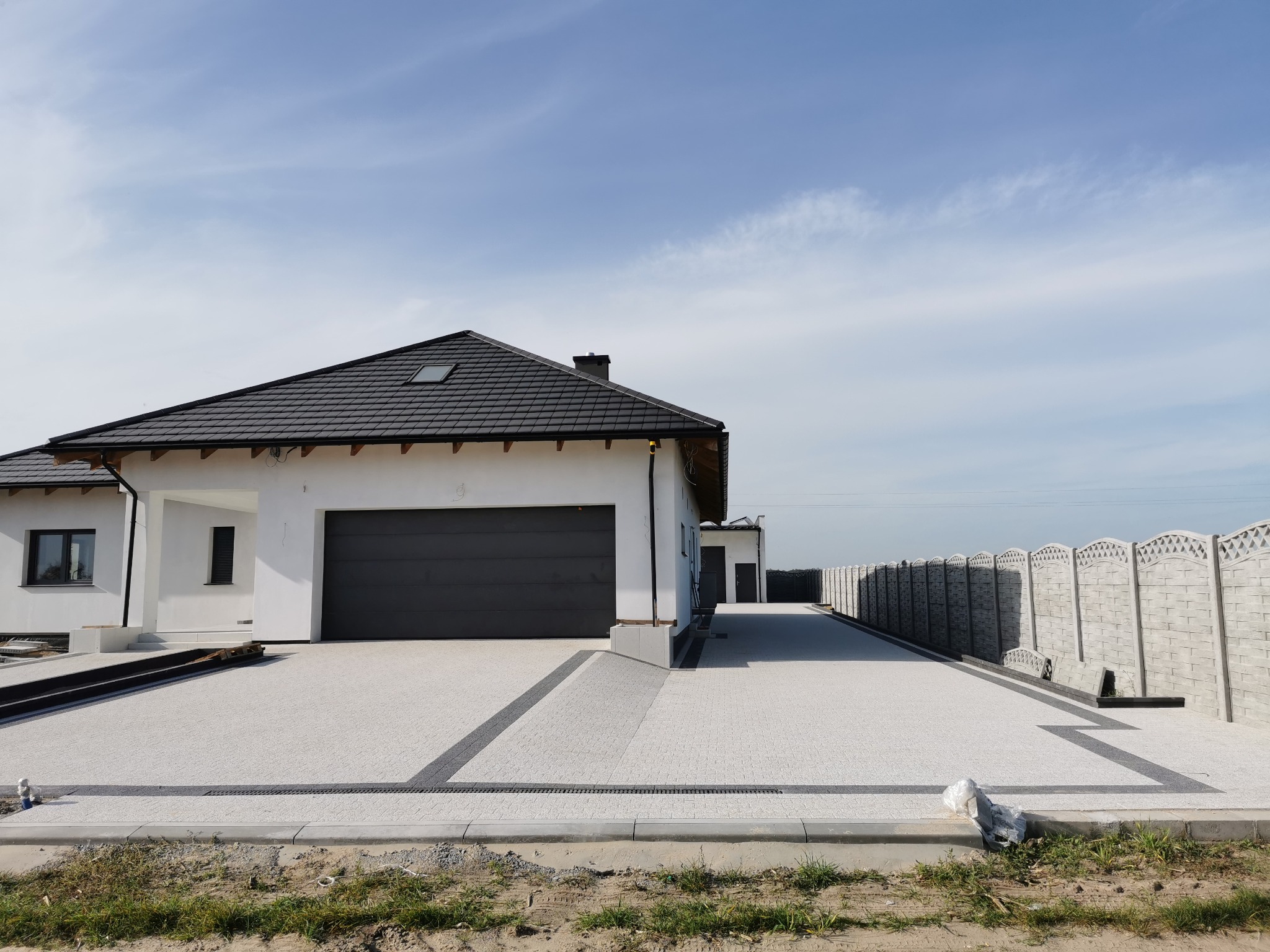 Nowoczesny dom jednorodzinny z jasną elewacją i czarnym dachem, podjazd wyłożony jasną kostką brukową z ciemnymi akcentami, widoczne studzienki odpływowe i fragment ogrodzenia.