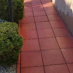 Czerwone płytki chodnikowe z radialnym wzorem, ułożone wzdłuż ściany budynku, obok przycięte krzewy i lampa ogrodowa