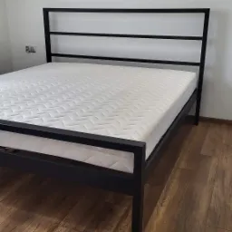 Minimalistyczne łóżko z czarną, metalową ramą i białym materacem w pokoju z drewnianą podłogą.