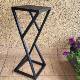 Czarny, metalowy kwietnik o geometrycznej konstrukcji, ustawiony na brązowych płytkach, z jasnym, kamiennym tłem i fioletowymi kwiatami w doniczce po prawej stronie.