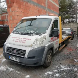 Biała laweta Fiat Ducato z numerem telefonu firmy na boku, częściowo pokryta śniegiem, przewozi szary samochód osobowy Renault Duster. W tle niedokończony budynek z czerwonej cegły i drzewa.