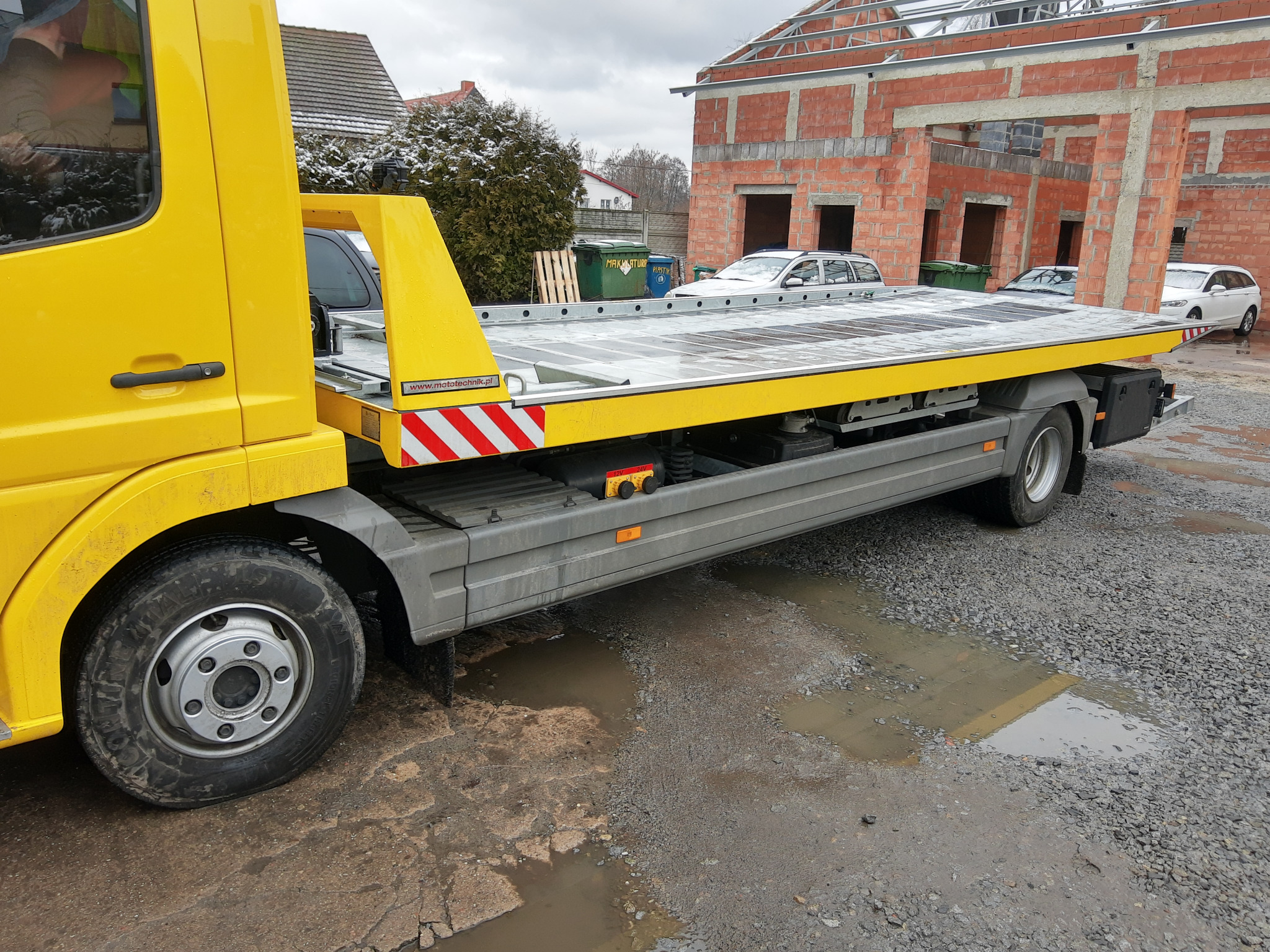 Żółta platforma transportowa z widocznym kołem i częścią kabiny, zaparkowana na żwirowym podłożu z kałużami, w tle budowa domu z czerwonej cegły i zaparkowane samochody.