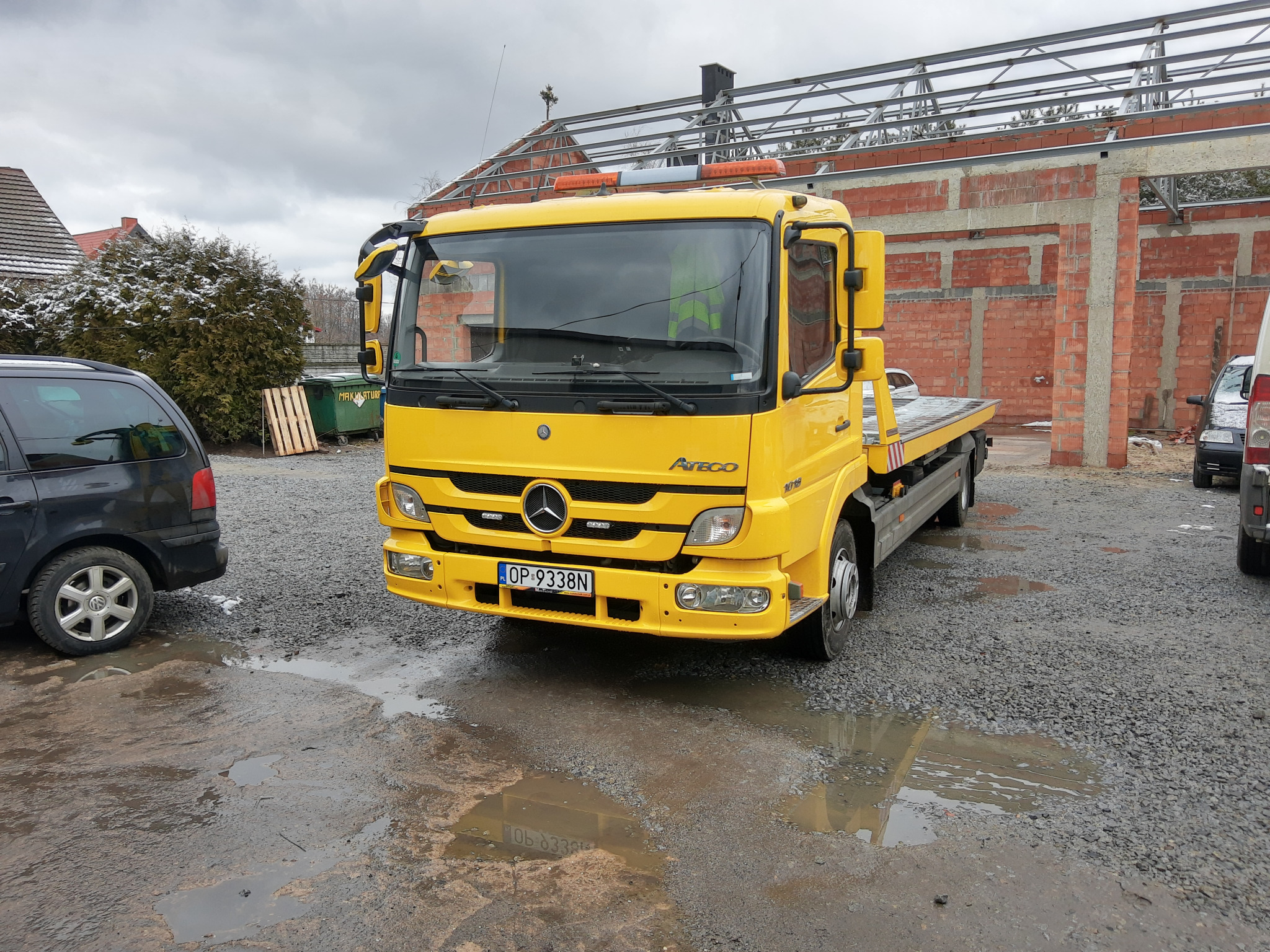 Żółta ciężarówka z platformą załadunkową marki Atego zaparkowana na placu budowy z kałużami, widoczne tablice rejestracyjne OP-9338N.