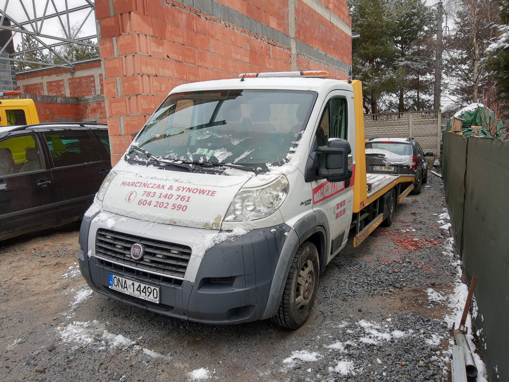 Biała laweta Fiat Ducato z numerem telefonu firmy na boku, częściowo pokryta śniegiem, przewozi szary samochód osobowy Renault Duster. W tle niedokończony budynek z czerwonej cegły i drzewa.