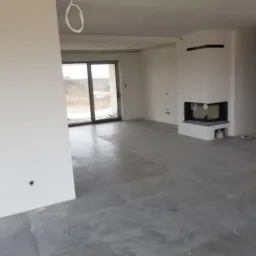 Surowe wnętrze z szarymi płytkami podłogowymi, białymi ścianami, widocznymi kablami i kominkiem narożnym w trakcie wykańczania.