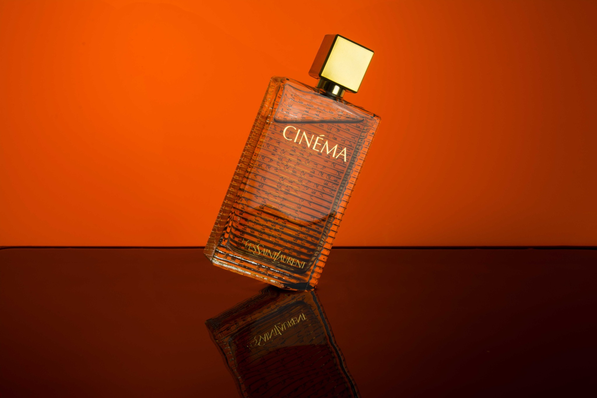 Butelka perfum Cinema Yves Saint Laurent na błyszczącym, ciemnym podłożu z pomarańczowym tłem, pochylona pod kątem.