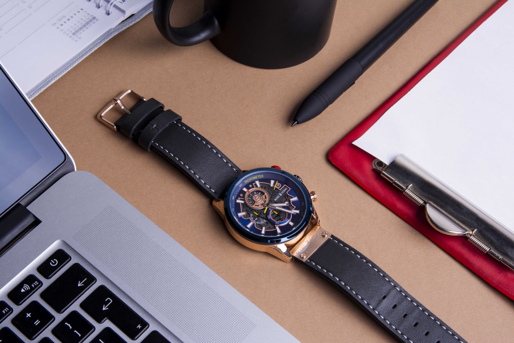 Zegarek męski na skórzanym pasku leżący na biurku obok laptopa, notatnika i czarnego długopisu.