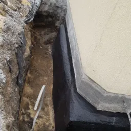 Fundament budynku zabezpieczony czarną, błyszczącą powłoką hydroizolacyjną, widoczny wykop w ziemi, fragment ściany budynku z jasnym tynkiem.