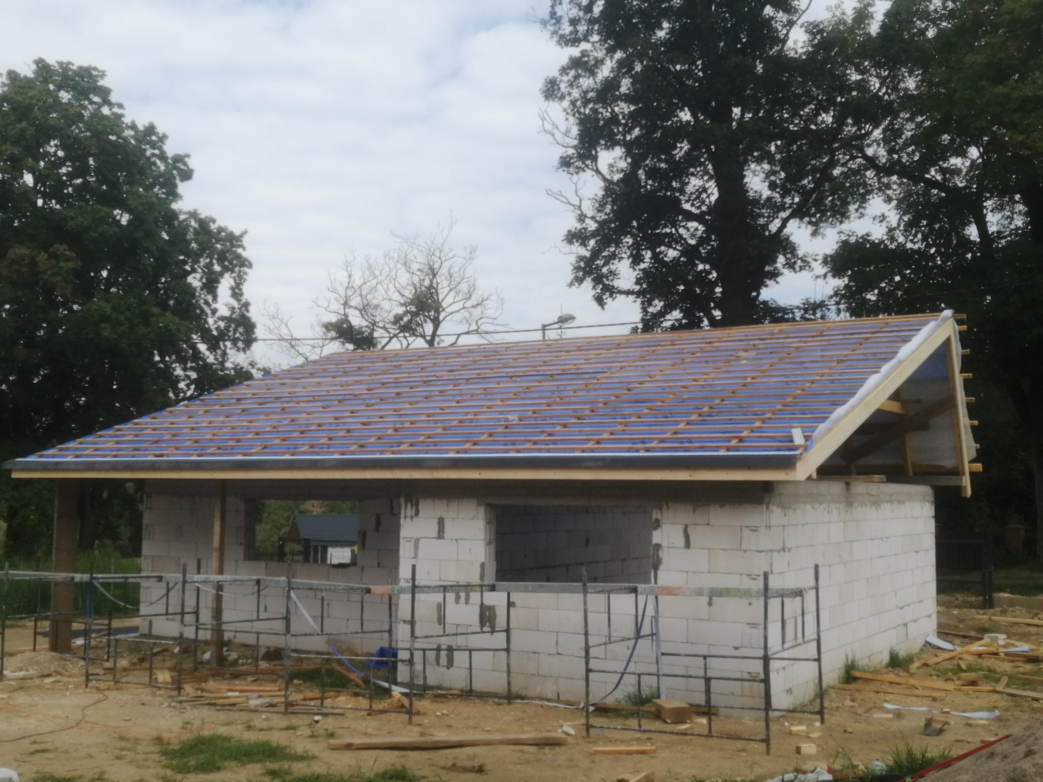 Wznoszony budynek z białych bloczków, z drewnianą konstrukcją dachu pokrytą niebieską folią, widoczne rusztowania i otoczenie drzew