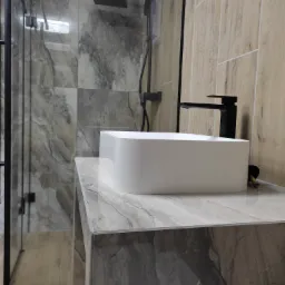 Nowoczesna łazienka z prostokątną, białą umywalką nablatową i czarną baterią, kabina prysznicowa ze szklaną ścianką i czarnym wykończeniem, płytki imitujące marmur na ścianach i blacie umywalki...