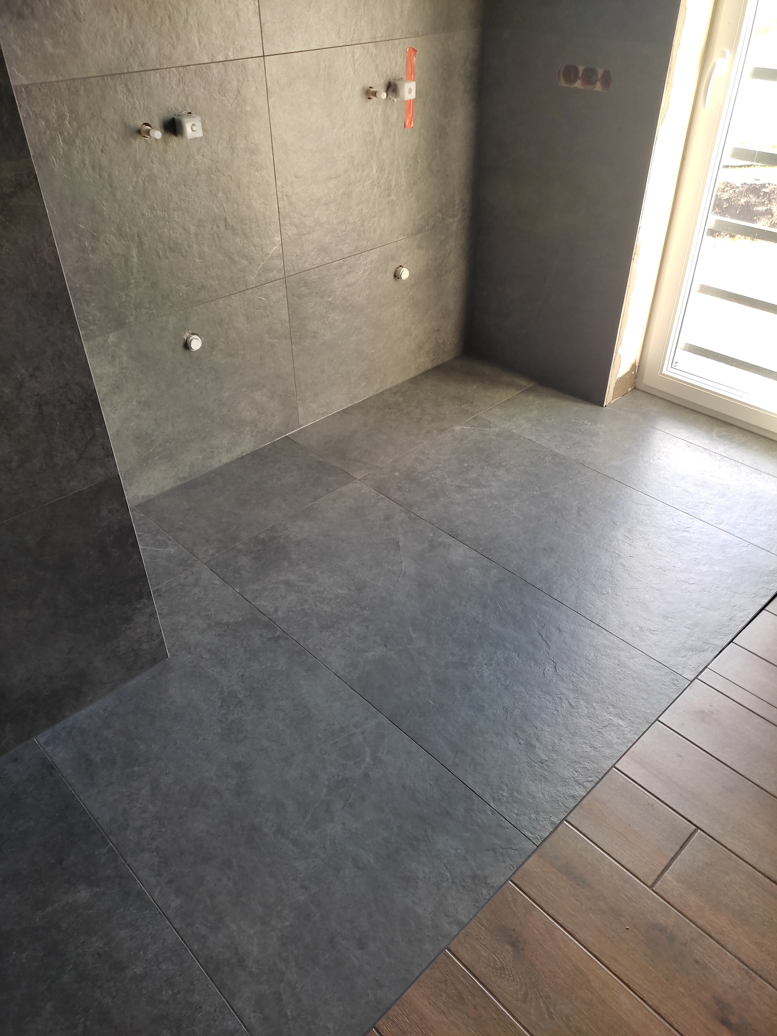 Ciemnoszare płytki imitujące kamień na ścianach i podłodze łazienki w trakcie prac wykończeniowych, widoczne wystające końcówki instalacji wodnej i elektrycznej, fragment drewnianej podłogi...