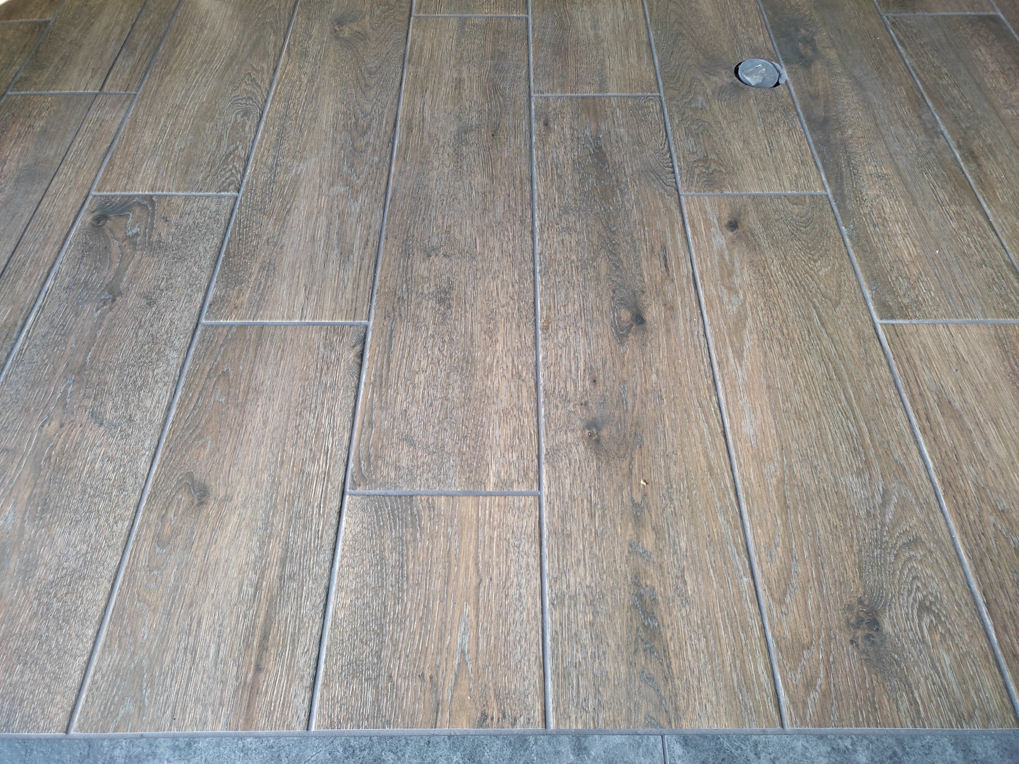 Ułożona pod kątem podłoga z płytek imitujących drewno z widocznym odpływem podłogowym i szarą listwą przypodłogową.