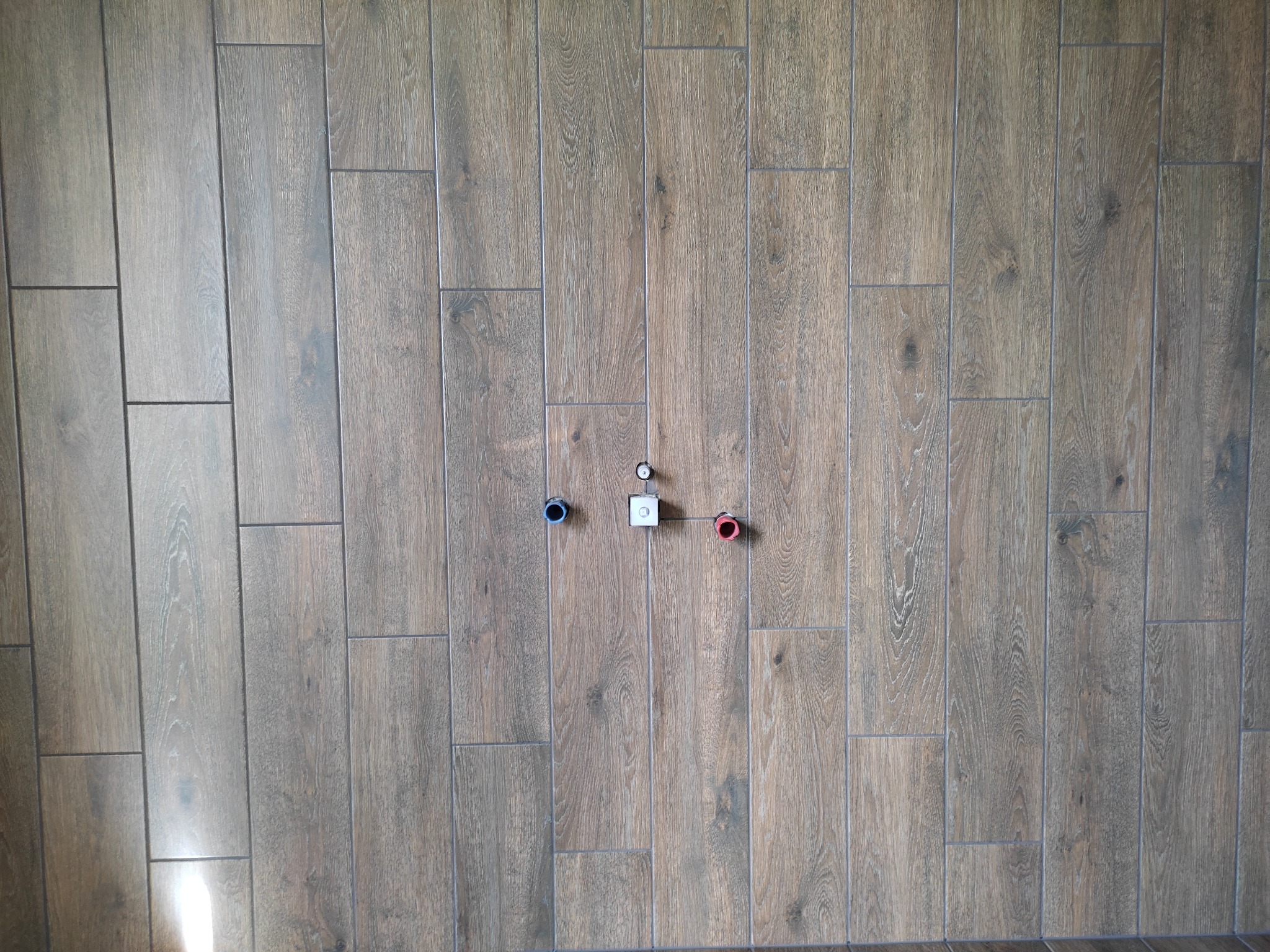 Ściana wyłożona płytkami imitującymi drewno w pionowym układzie z widocznymi przyłączami wodnymi: zimnej (niebieskie oznaczenie), ciepłej (czerwone oznaczenie) i odpływu, przygotowana do montażu...