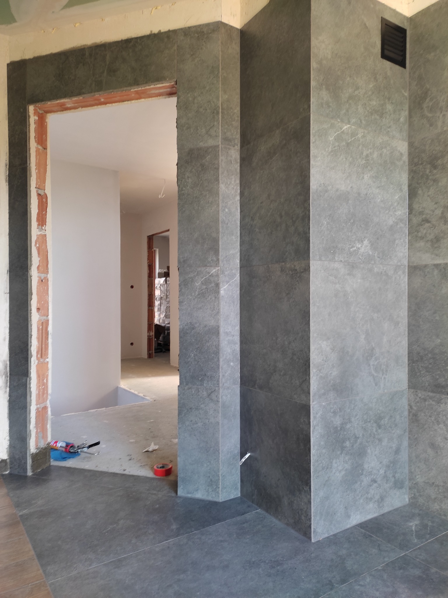 Montaż szarych płytek imitujących kamień na ścianie i podłodze, widoczny fragment futryny drzwiowej z cegły oraz narzędzia pracy.