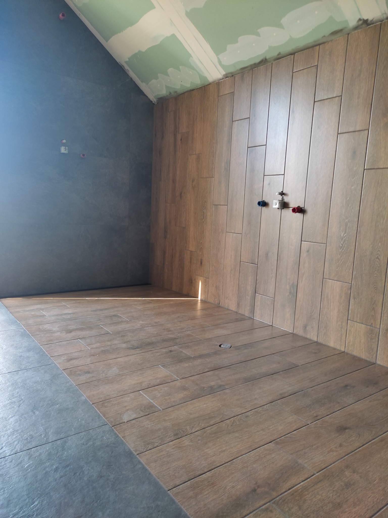 Wnętrze łazienki w trakcie prac wykończeniowych: położona podłoga i ściana w płytkach imitujących drewno, surowa ściana w szarym kolorze, widoczne przyłącza wodne.