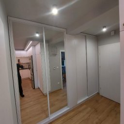 M&A DESIGN - Biała szafa wnękowa z przesuwnymi drzwiami z lustrami, odbicie kuchni w lustrach, podłoga z paneli drewnopodobnych.