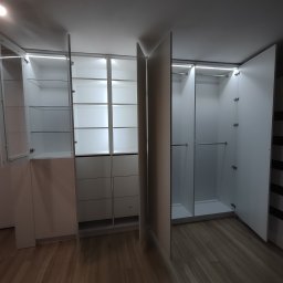 M&A DESIGN - Biała szafa wnękowa z oświetleniem LED, częściowo otwarta, z widocznymi półkami szklanymi i szufladami, na drewnianej podłodze.