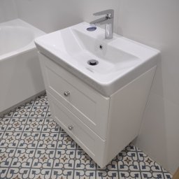 M&A DESIGN - Biała umywalka z szafką z dwiema szufladami i chromowaną armaturą, na tle białych płytek ściennych i wzorzystej podłogi.