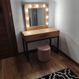 M&A DESIGN - Drewniana toaletka z lustrem podświetlanym żarówkami, z szufladą i czarnymi metalowymi nogami, obok różowy puf, na podłodze drewniane panele i fragment czarno-białego dywanu.