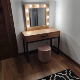 Drewniana toaletka z lustrem podświetlanym żarówkami, z szufladą i czarnymi metalowymi nogami, obok różowy puf, na podłodze drewniane panele i fragment czarno-białego dywanu.