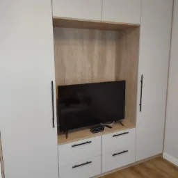 Biała szafa na wymiar z wnęką na telewizor i szufladami, czarne uchwyty, drewnopodobne wykończenie wnęki, widoczny parkiet