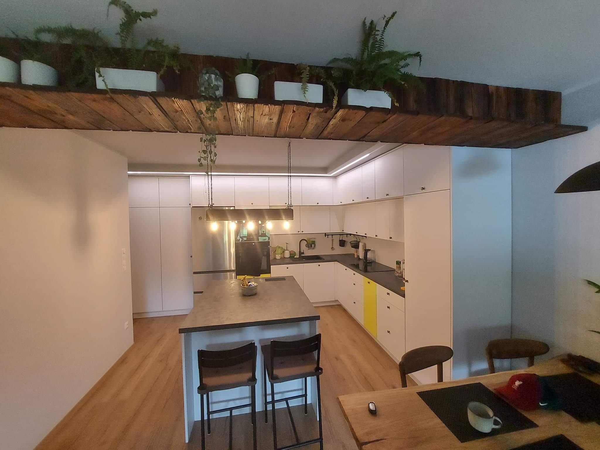 Nowoczesna kuchnia z białymi szafkami, wyspą z czarnym blatem i drewnianym sufitem z roślinami doniczkowymi, oświetlona wiszącymi lampami.