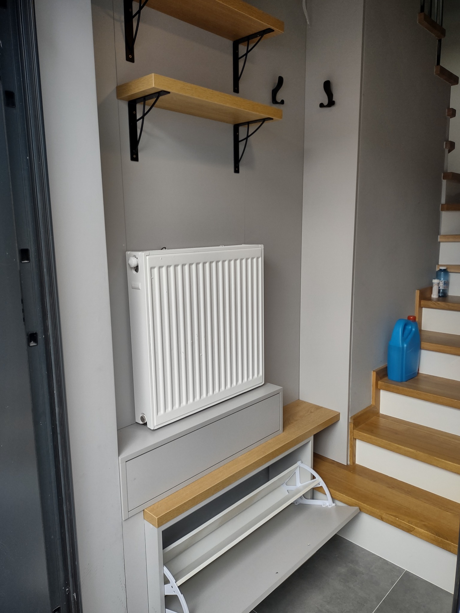 Szara zabudowa przedpokoju z grzejnikiem, półkami na wspornikach i szafką na buty z uchylnym frontem, widoczne schody z drewnianymi stopniami.