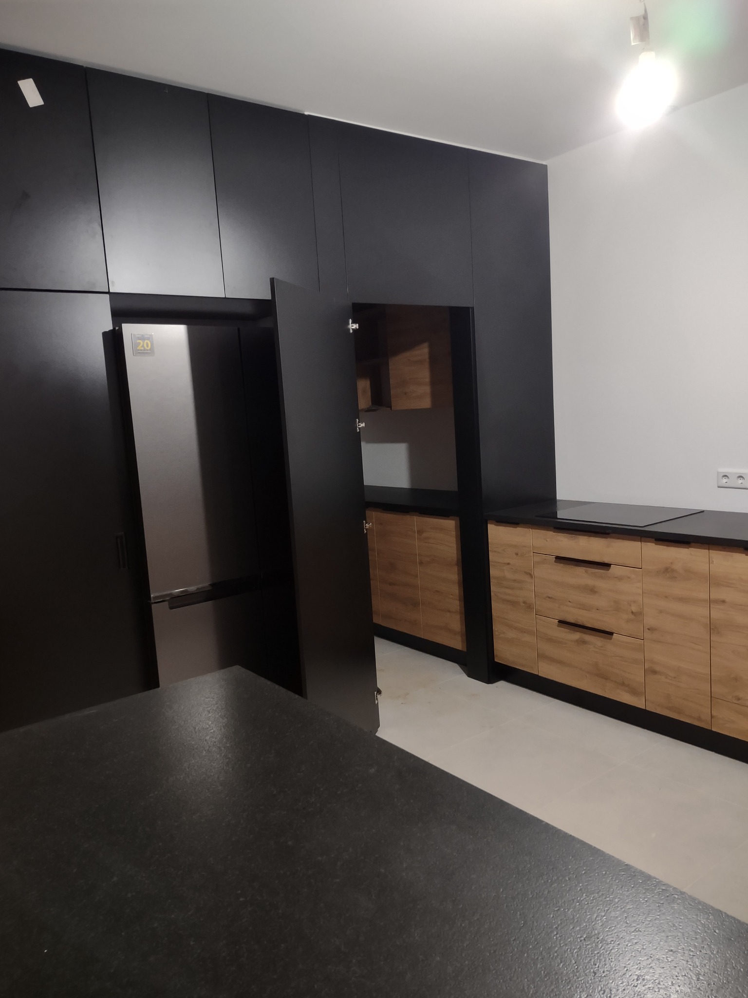 Nowoczesna kuchnia z czarnymi szafkami górnymi i drewnopodobnymi szafkami dolnymi, z otwartym frontem szafki odsłaniającym wnętrze, widoczna lodówka w zabudowie i czarny blat.