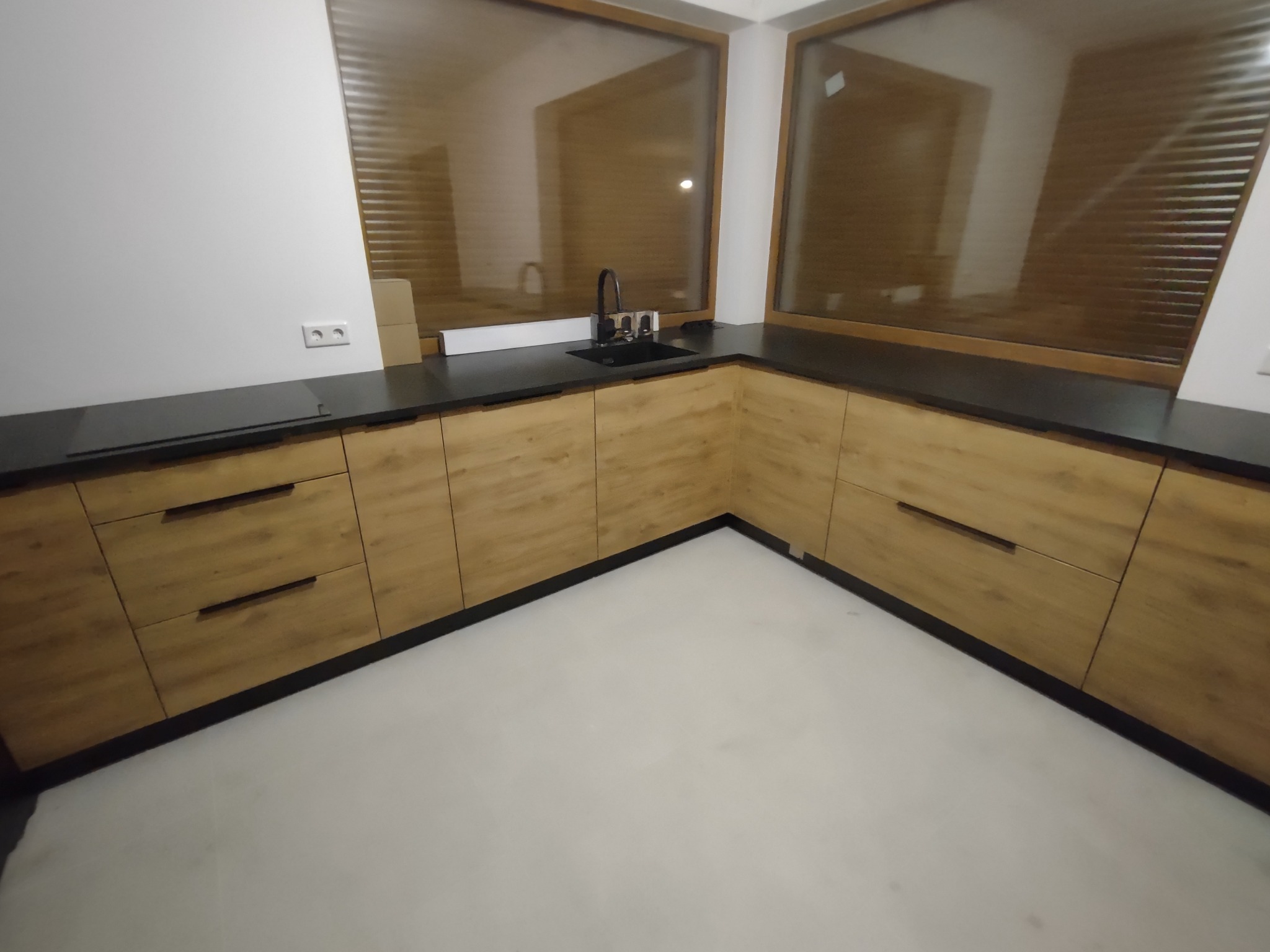 Kąt ujęcia na nowoczesną kuchnię z drewnianymi frontami szafek, czarnym blatem i oknami z żaluzjami w tle.