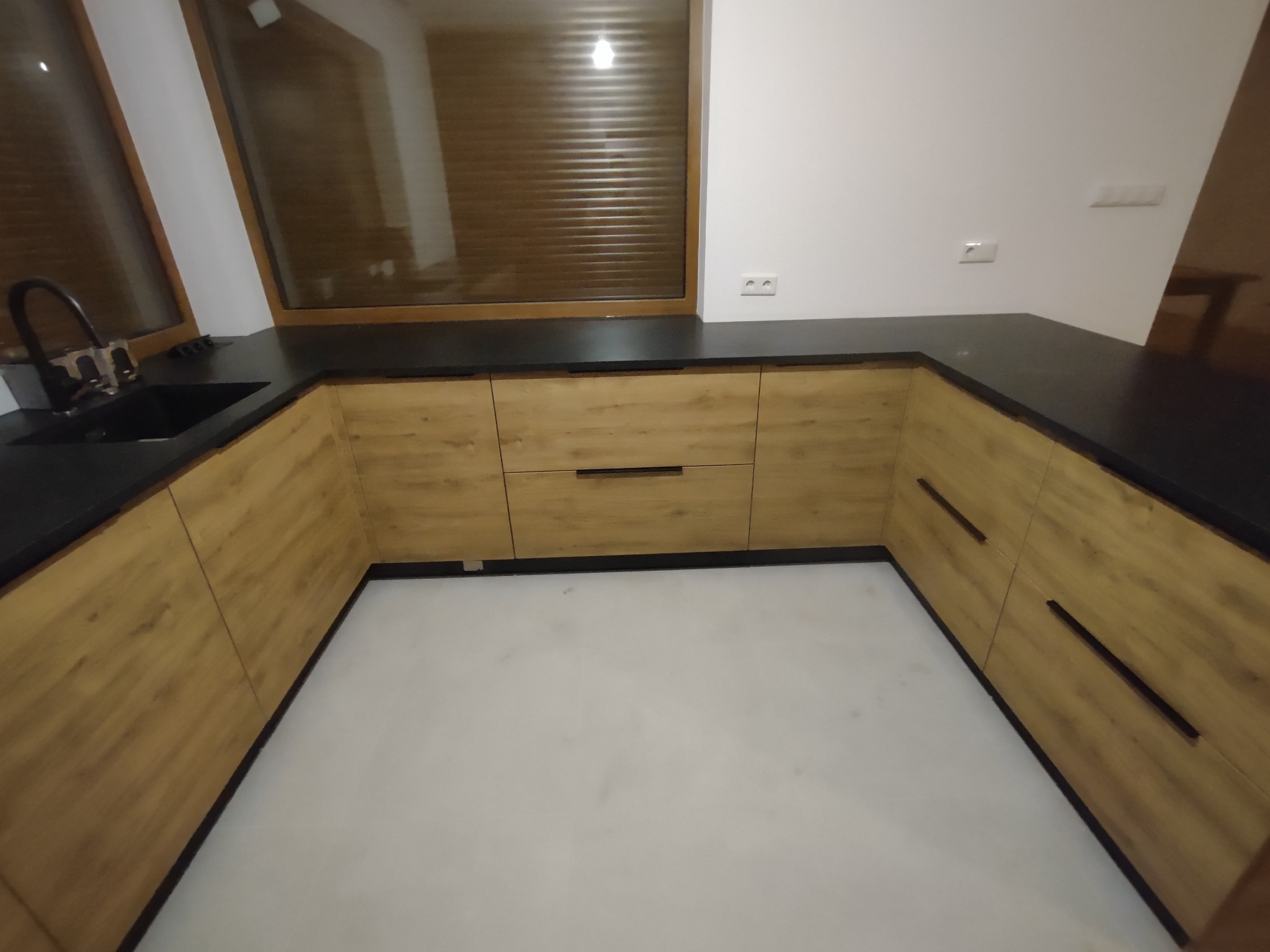 Widok na narożną zabudowę kuchenną z drewnianymi frontami i czarnym blatem, okno z brązowymi roletami w tle, widoczny czarny zlew z baterią.